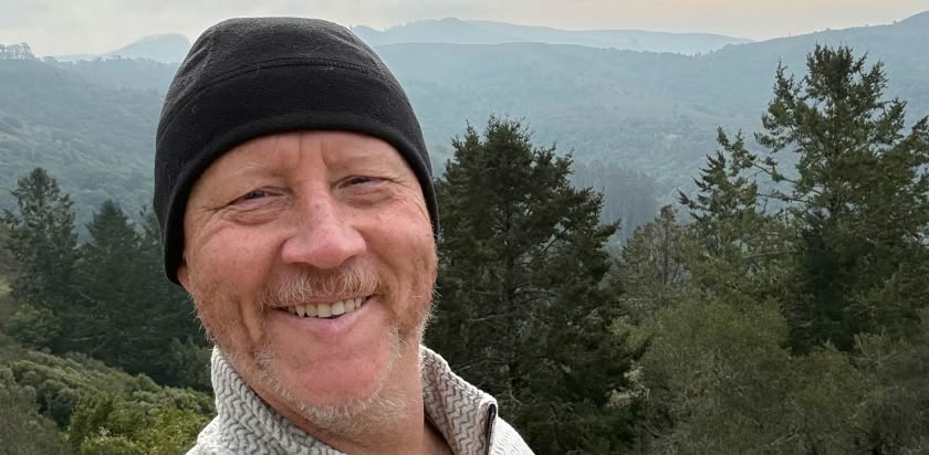 John Ogle takes a selfie while enjoying an active lifestyle after cancer treatment. Hills and trees are in the background and mountains are in the distance 