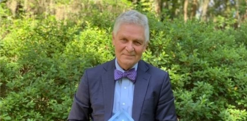 Dr. William “Bill” Mendenhall in an outdoor setting wearing a suit and bowtie on the occasion of receiving in 2020 a lifetime achievement award, which he holds in his hands.