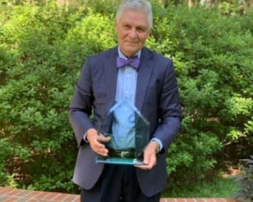 Dr. William “Bill” Mendenhall in an outdoor setting wearing a suit and bowtie on the occasion of receiving in 2020 a lifetime achievement award, which he holds in his hands.