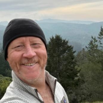 John Ogle takes a selfie while enjoying an active lifestyle after cancer treatment. Hills and trees are in the background and mountains are in the distance 