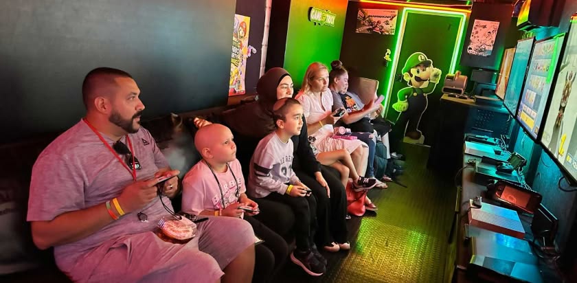 a group of parents and children are seated on bench inside a mobile video game truck and are intently looking at the screens on the opposite wall while holding video game controls