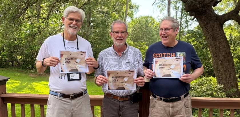 three men smiling and standing together on a wooden deck with a grassy lawn and trees in the background, each holding a photo of a proton treatment room printed with their names and the dates they were treated